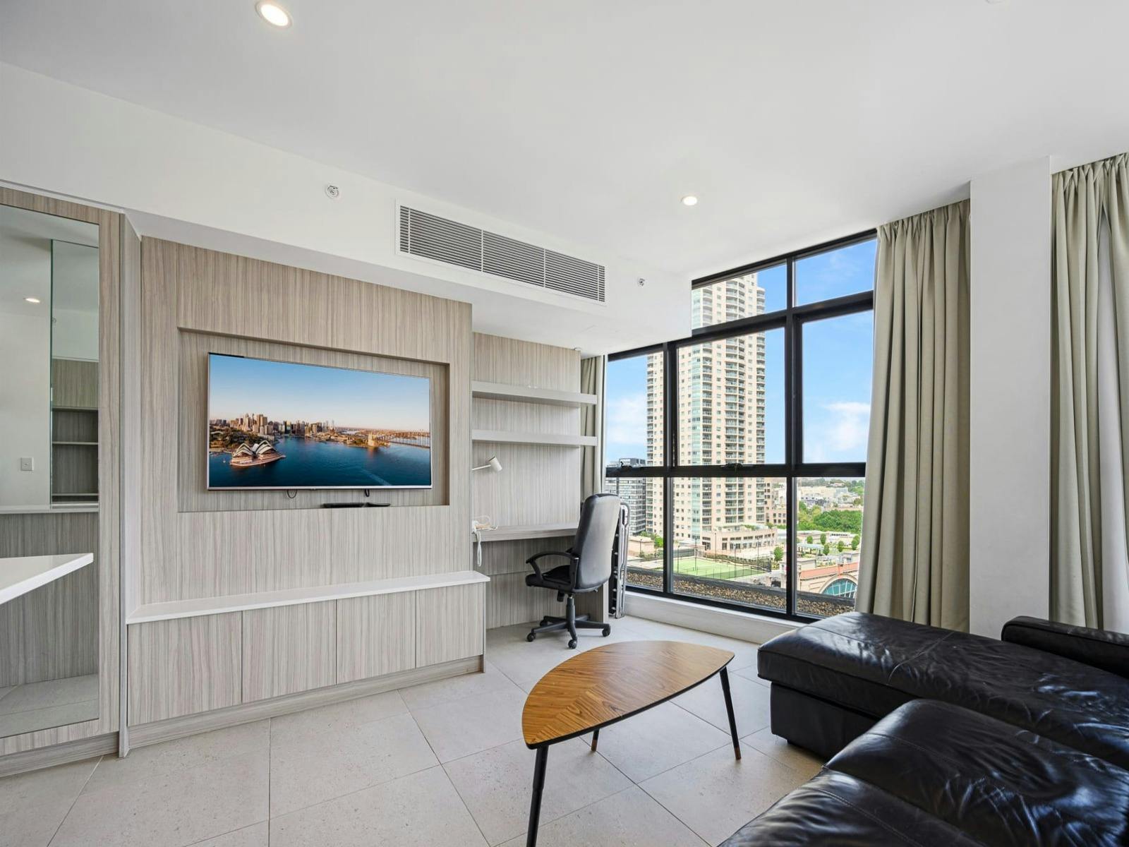 Living room with leather sofa seating, wall-mounted TV, study desk, and large windows with city view