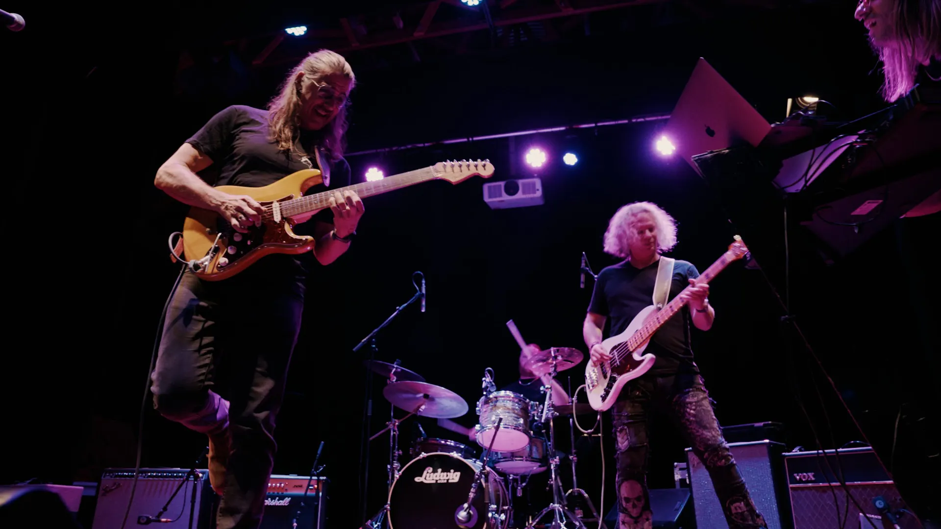 Two men on a stage playing guitar, dark background with three purple lights