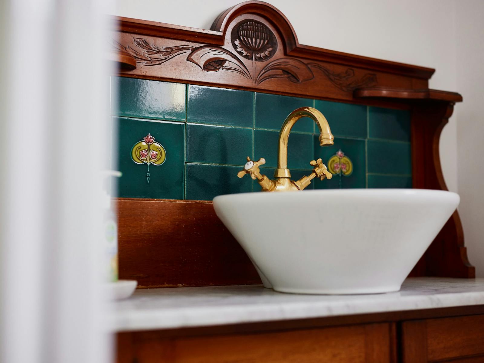 A basin sits on a darn, wooden vanity with deep green tiles. The tap ware is shiny and gold.
