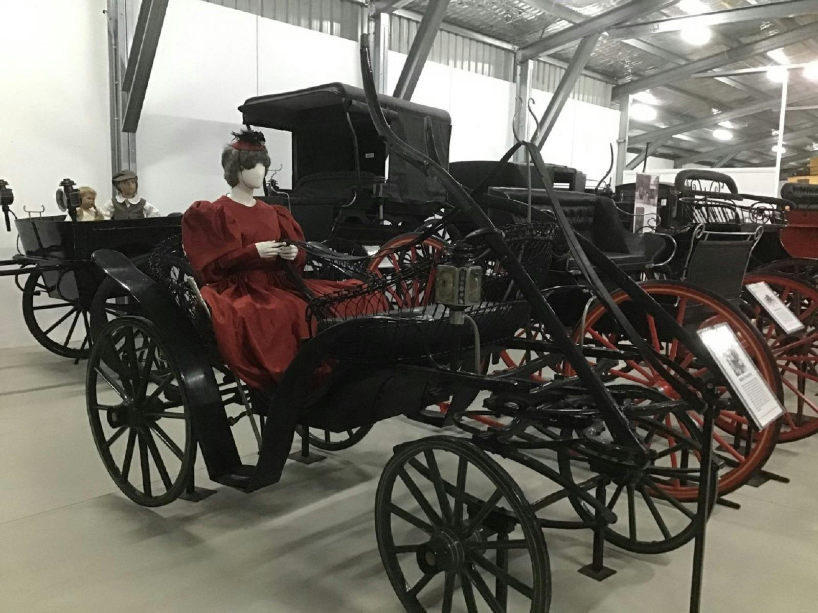 A beautifully restored black, old horse drawn buggy with a lady dressed in a red gown and a hat.
