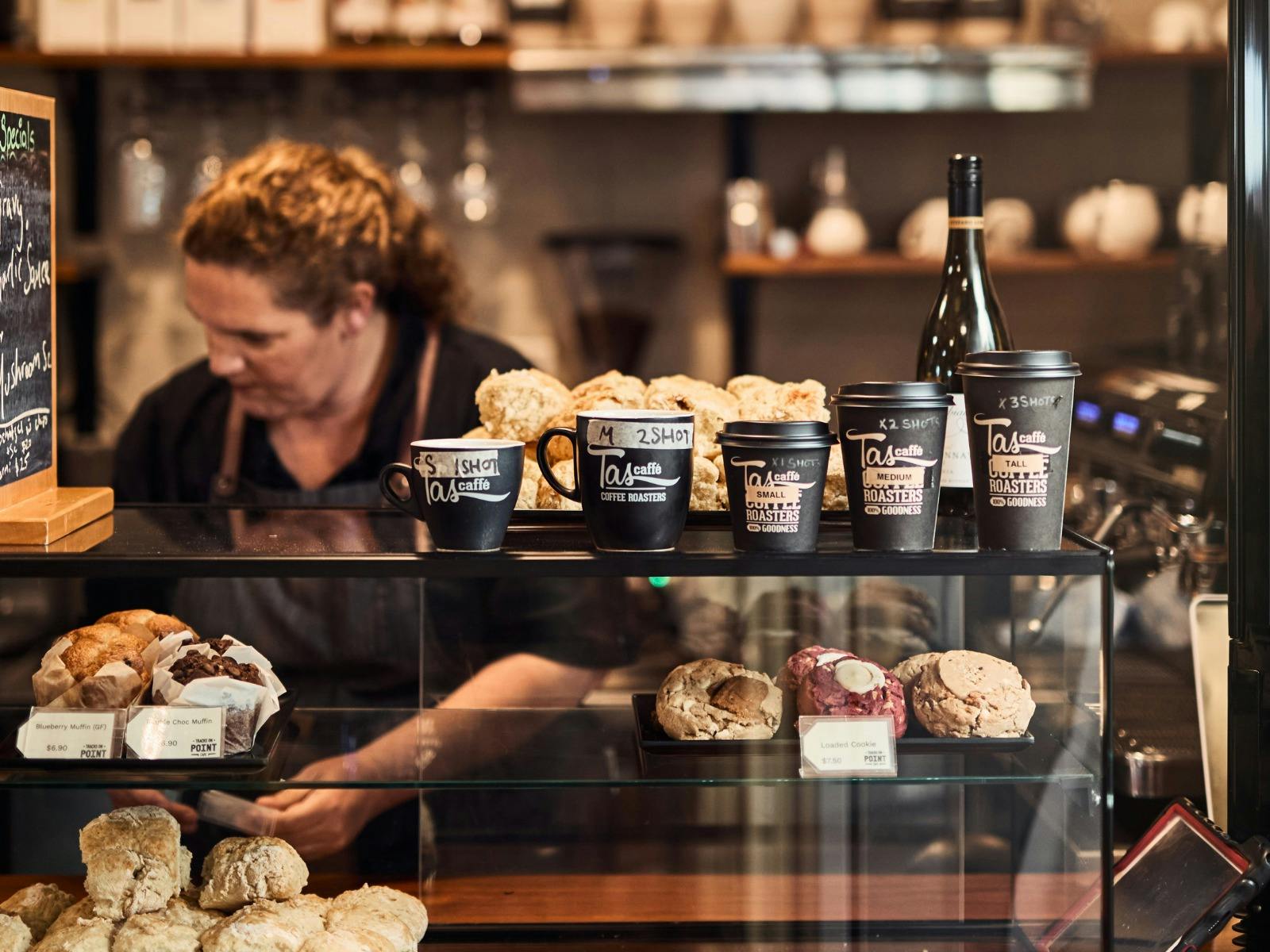 Woman in a black tshirt and apron works behind a cafe counter with coffee cups and biscuits on it