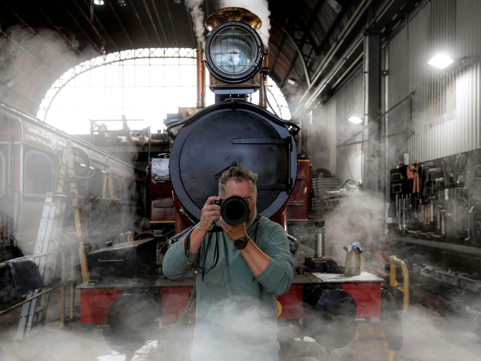 Cam Blake faces the camera with a DSLR camera held to his face. Behind him is a steam locomotive.