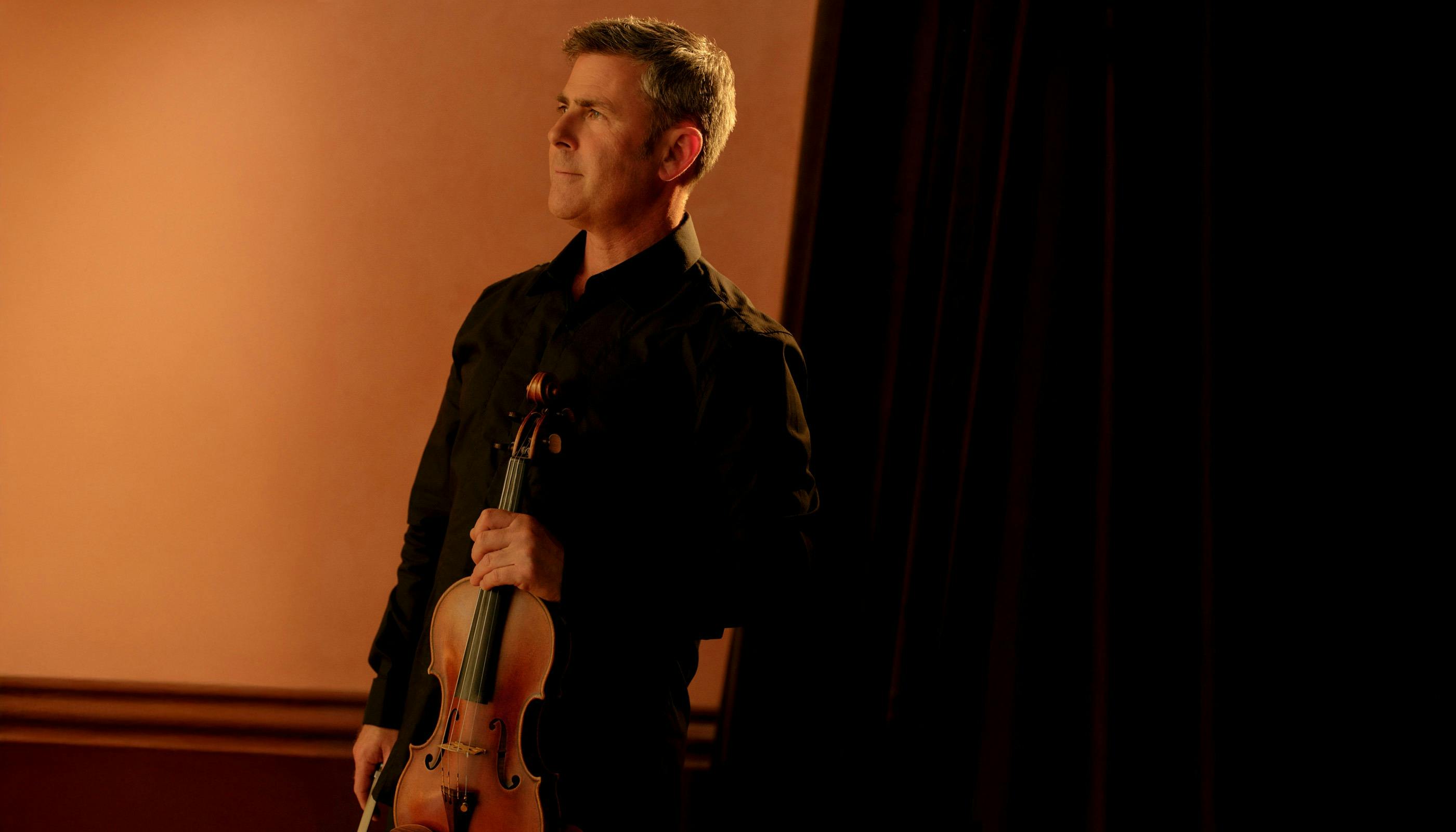 A Man in black holds a violin and stares off camera to the left