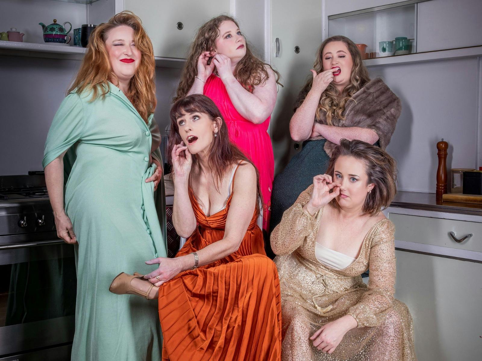 Five women in colourful dresses strike different poses against the backdrop of a kitchen