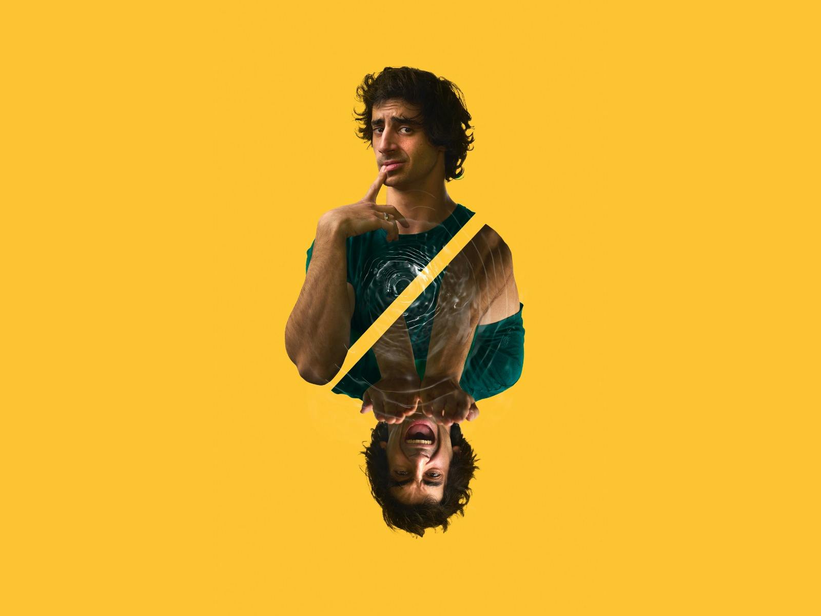 A man with curly hair stands against a yellow background, with his finger on his lips