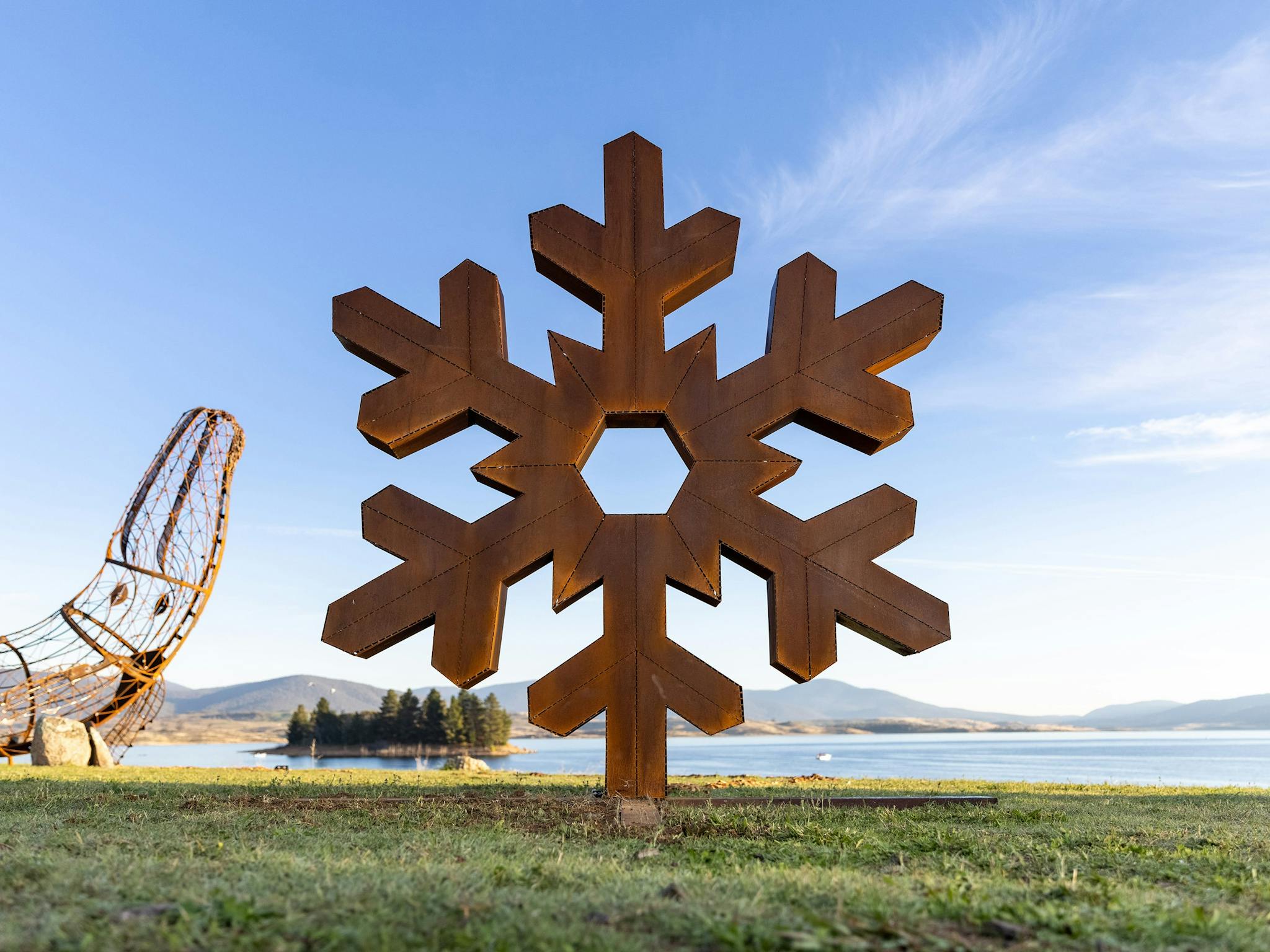 A large metal sculpture of a snowflake, set on green grass above a clear lake and blue sky backdrop.