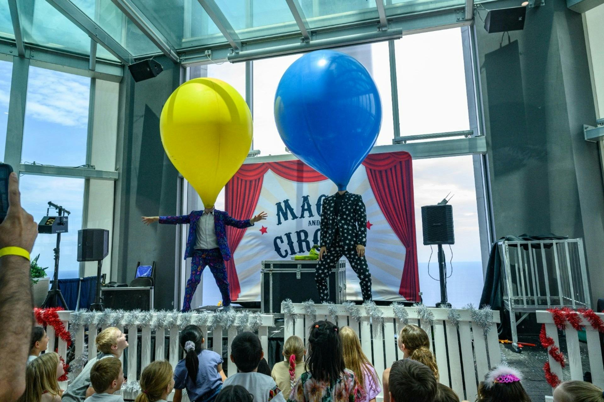 Two performers on stage with giant yellow and blue balloons while children watch from the audience