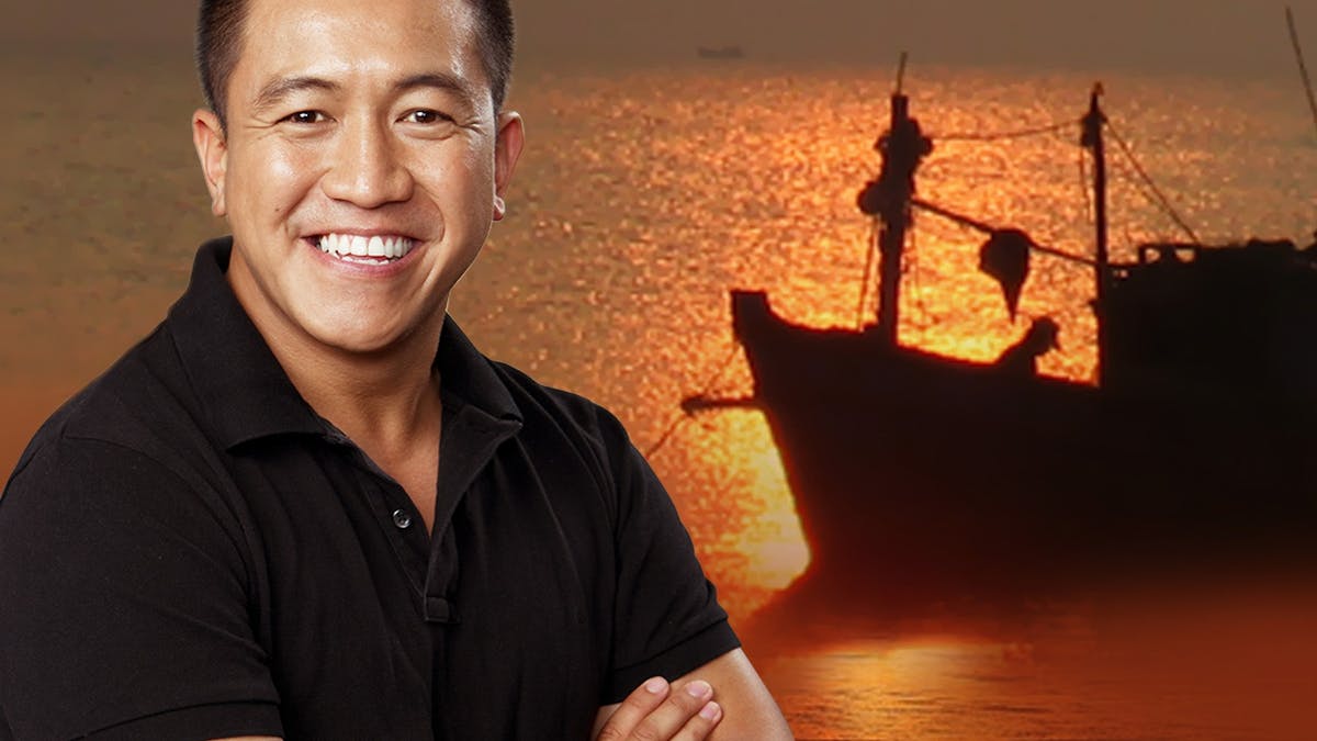 An Asian man in a black tshirt standing in front of a background of the ocean and boat in silhouette