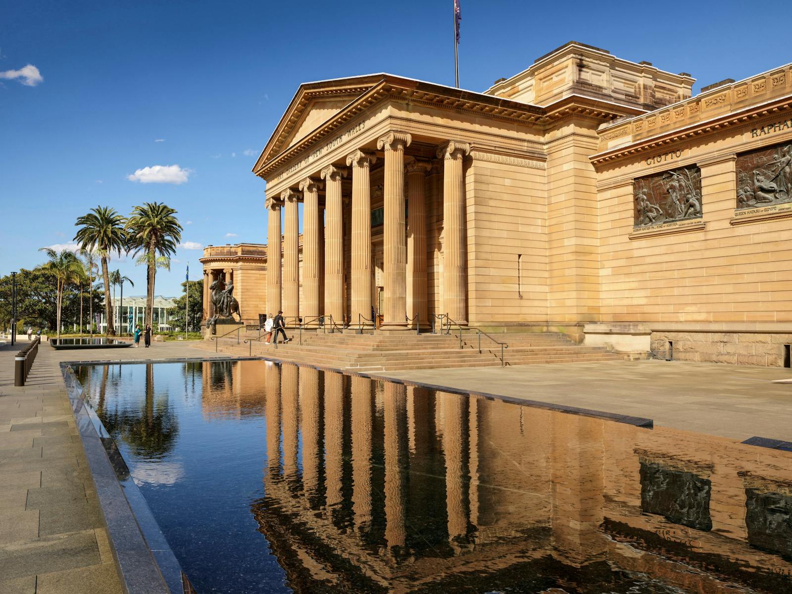 Grand sandstone building with tall columns, reflected in a clear rectangular pool