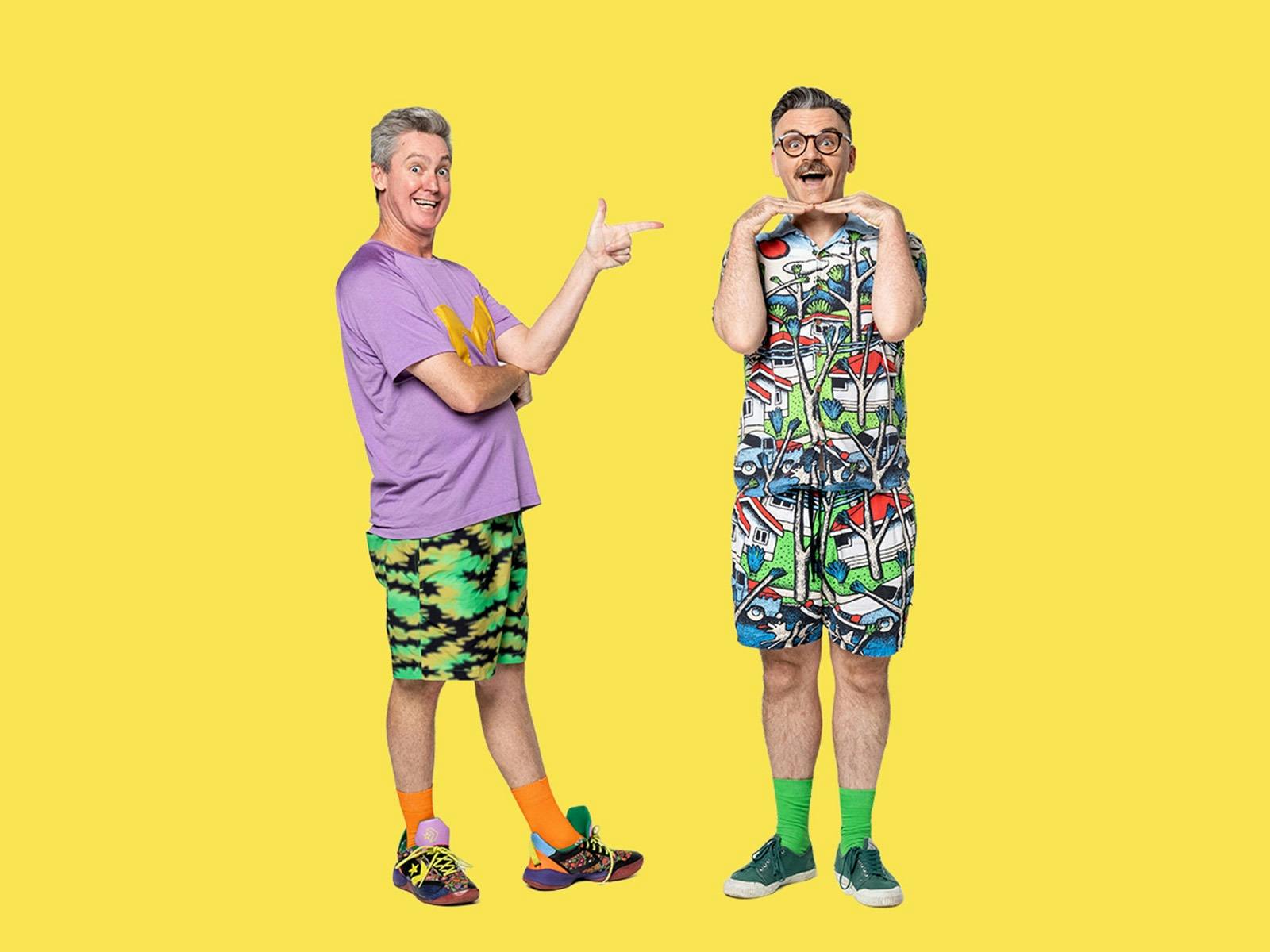 Two men in colorful, playful outfits pose against a yellow background, each tilting their head