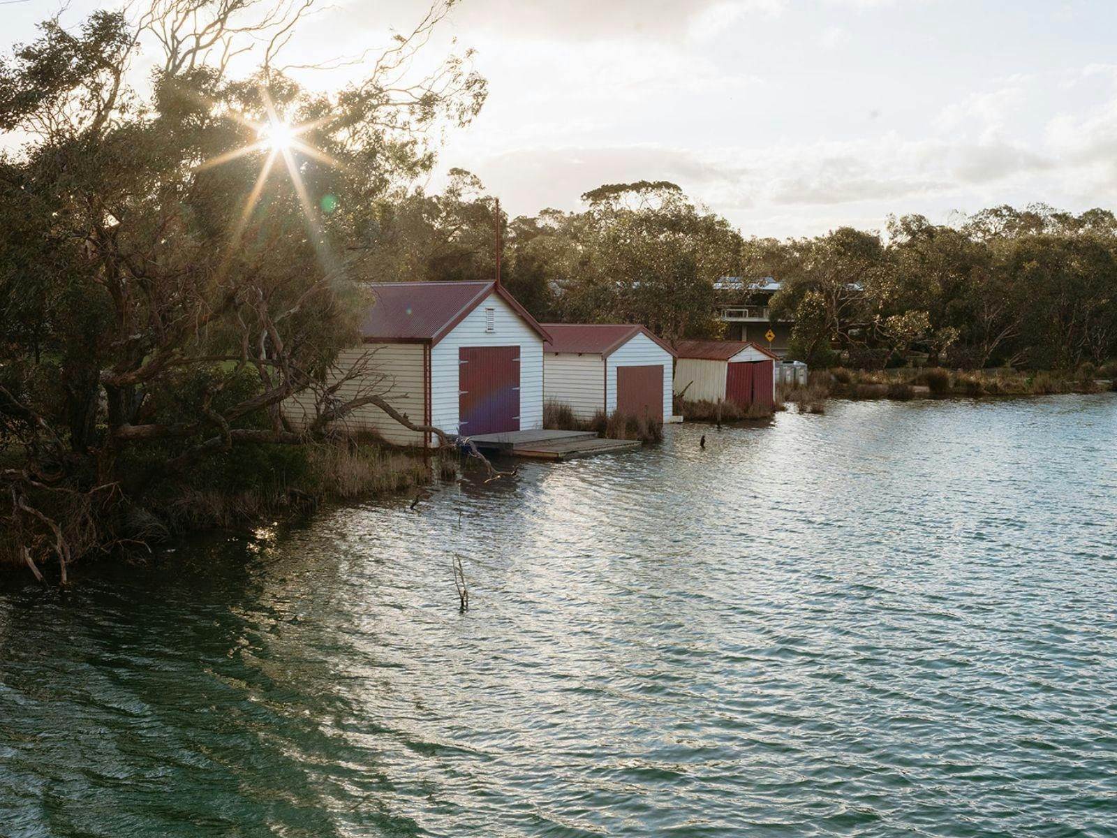 Boathouses along a river at sunset with sunlight through trees and gentle water reflections.