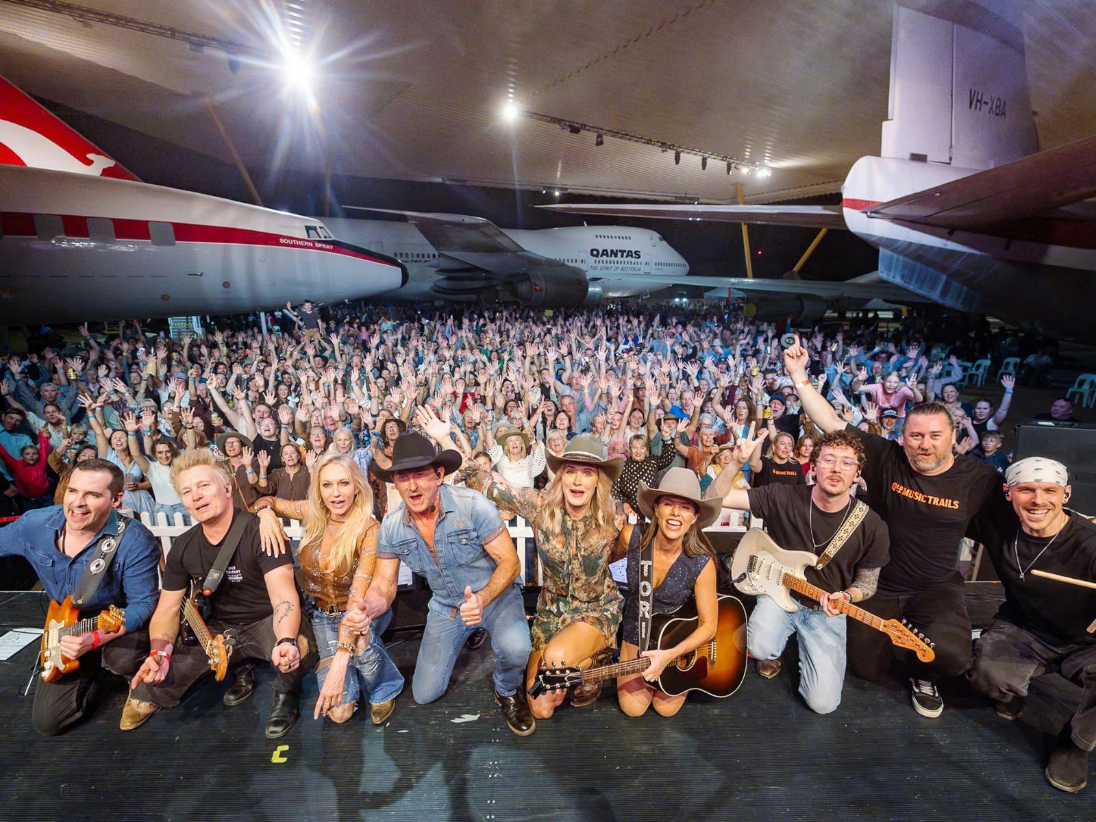 Musicians on stage posing for the camera with a large crowd cheering in the Qantas Founders Museum