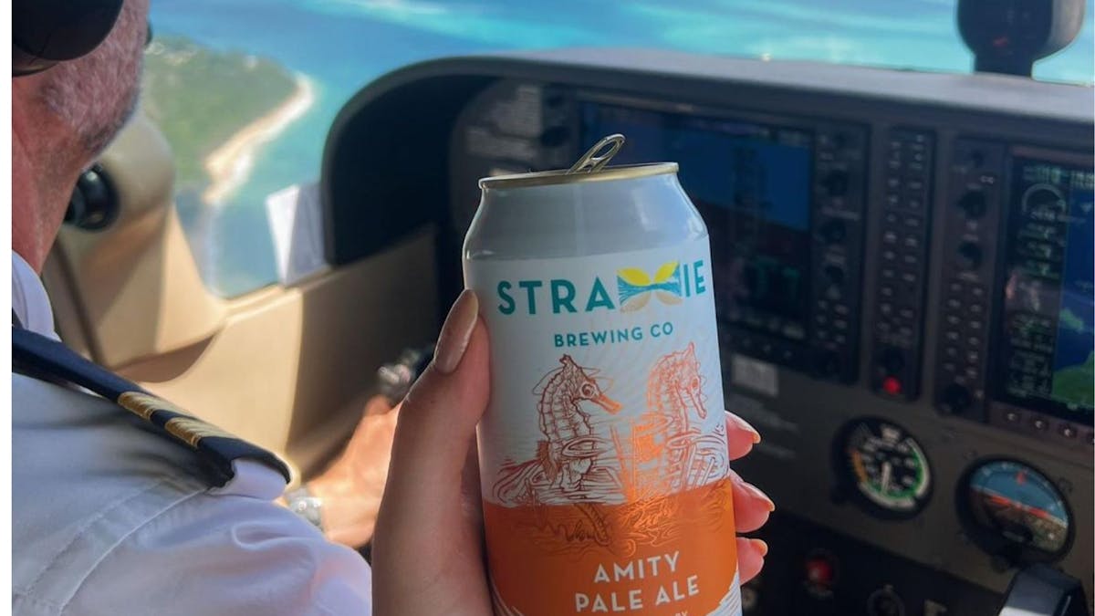 Hand holding a can of Amity Pale Ale beer in foreground. Background is interior of plane and pilot