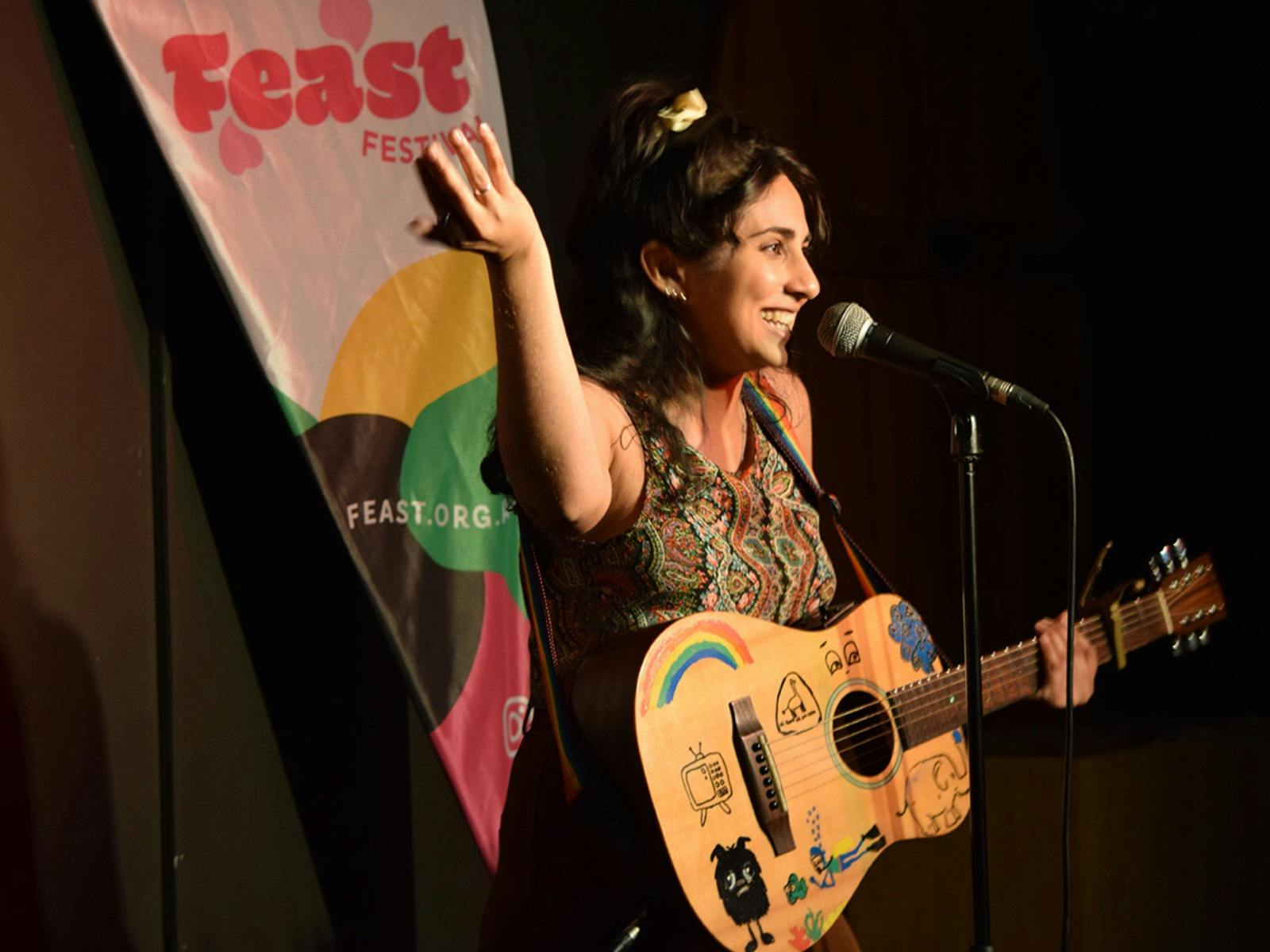 A performer standing by the mic waving her hand in the air and holding a guitar with cartoon doodles