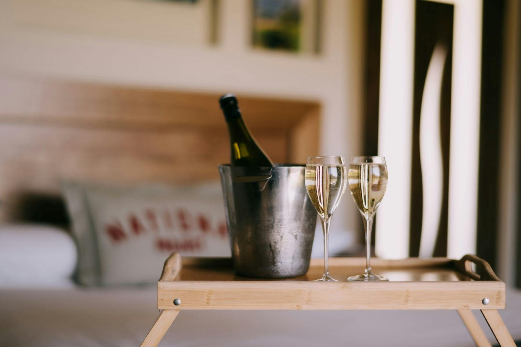 A wooden tray with champagne glasses and a chilled bottle in an ice bucket on a cozy bed.