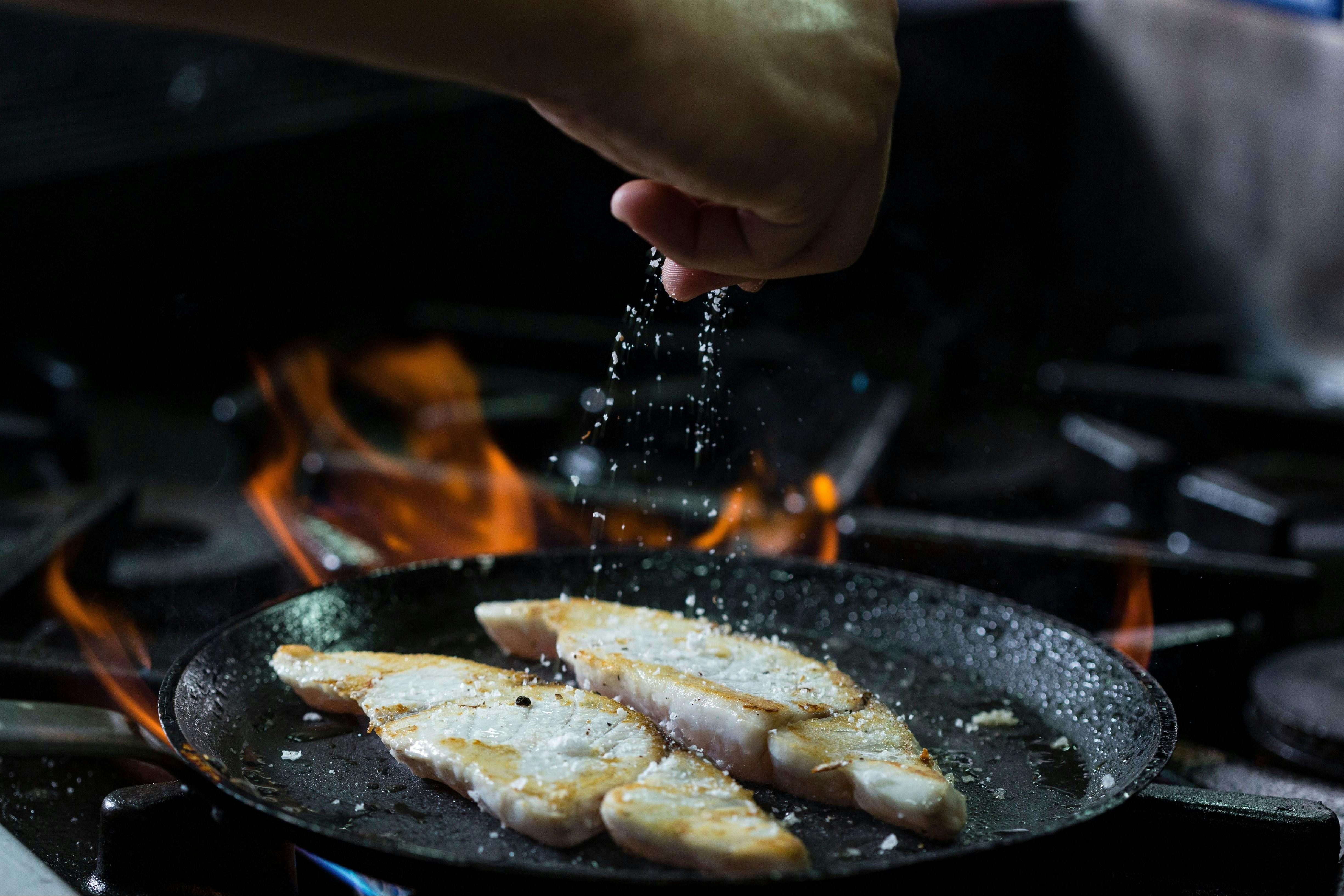 A chef seasoning fresh fish fillets in a flaming cast iron pan.