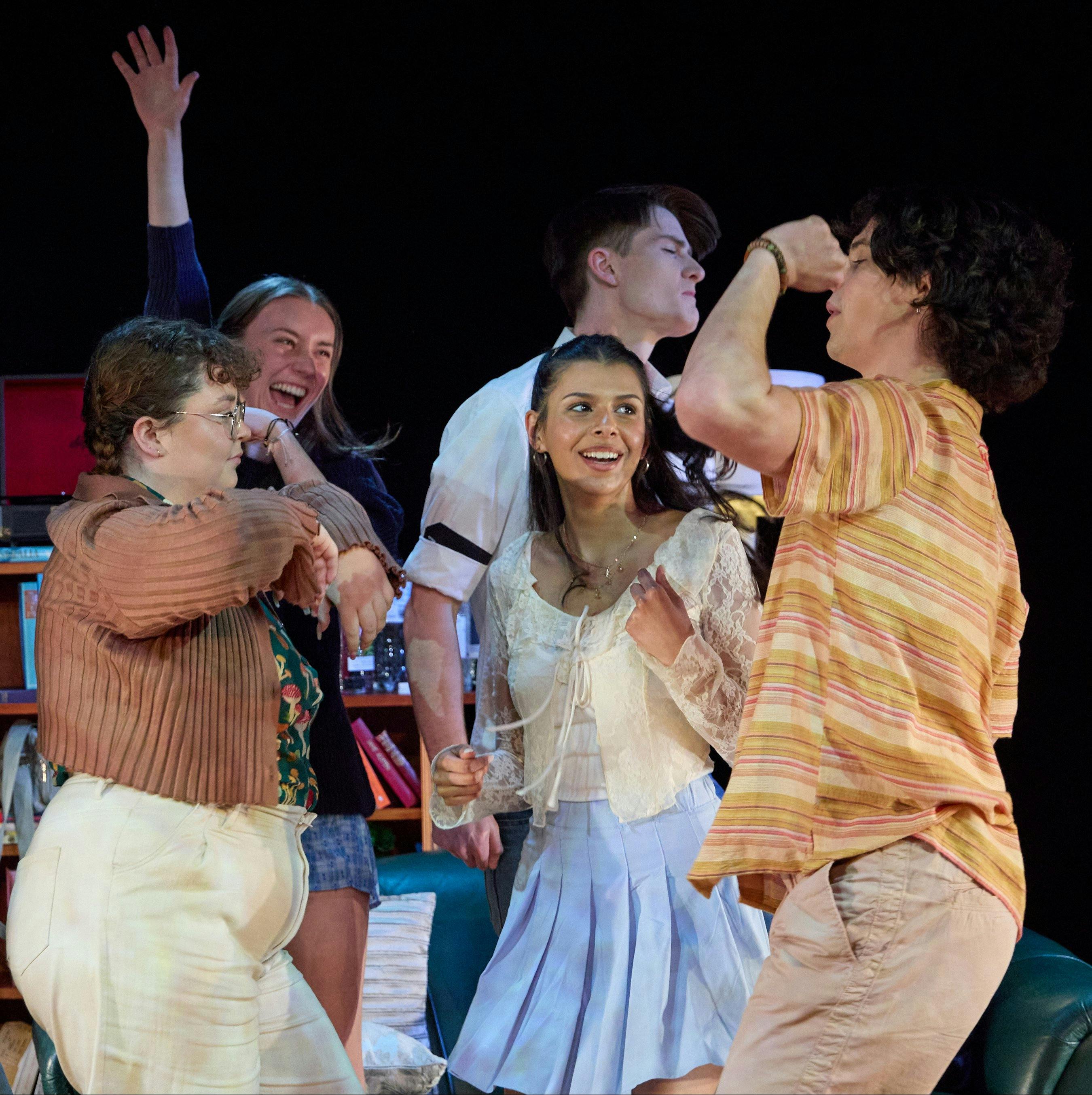 A group of teenagers dance wildly in a stage set of a living room in a theatre production.