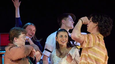 A group of teenagers dance wildly in a stage set of a living room in a theatre production.