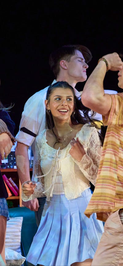 A group of teenagers dance wildly in a stage set of a living room in a theatre production.