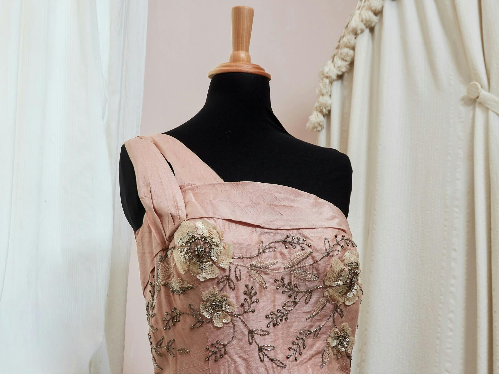 Stunning one shoulder light pink dress with stitched embellishments sits upon a black mannequin