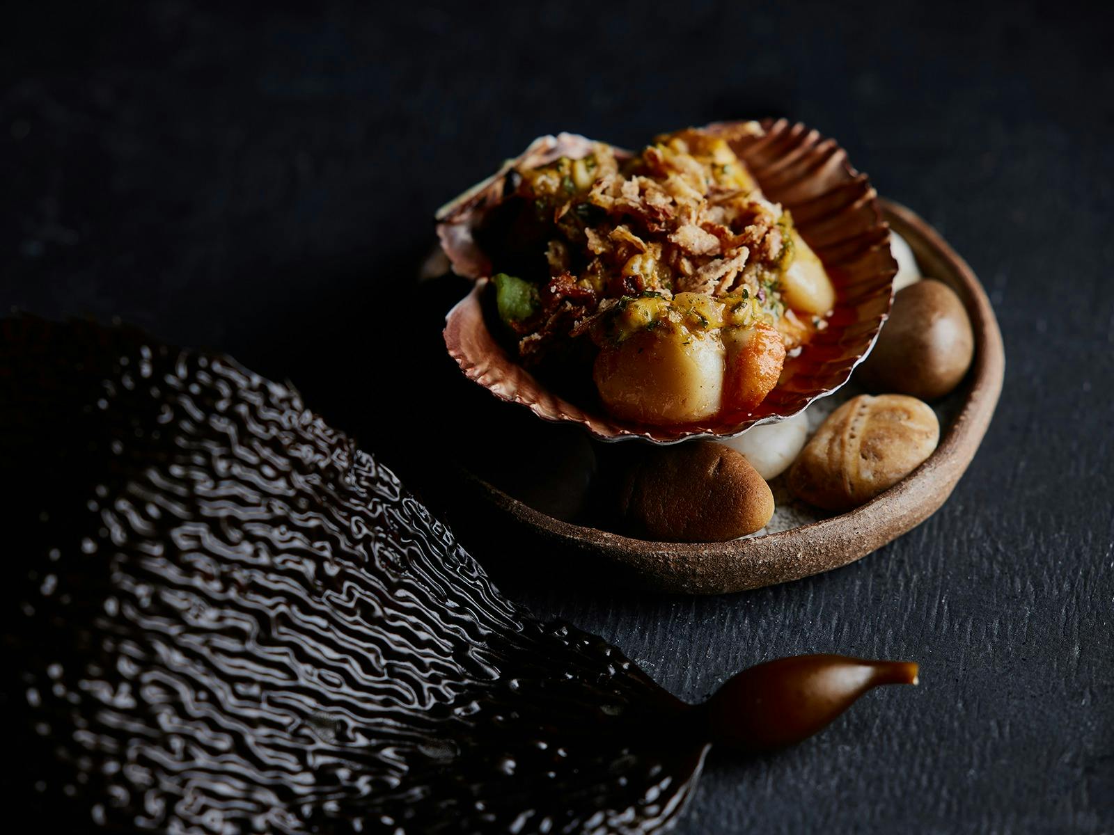 Scallops served in a shell, presented on a small plate with rocks and a piece of seaweed.