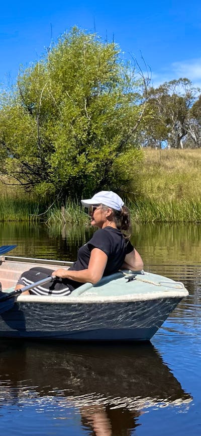 Man, woman & dog in small rowboat on water, blue sky, grass & trees behind