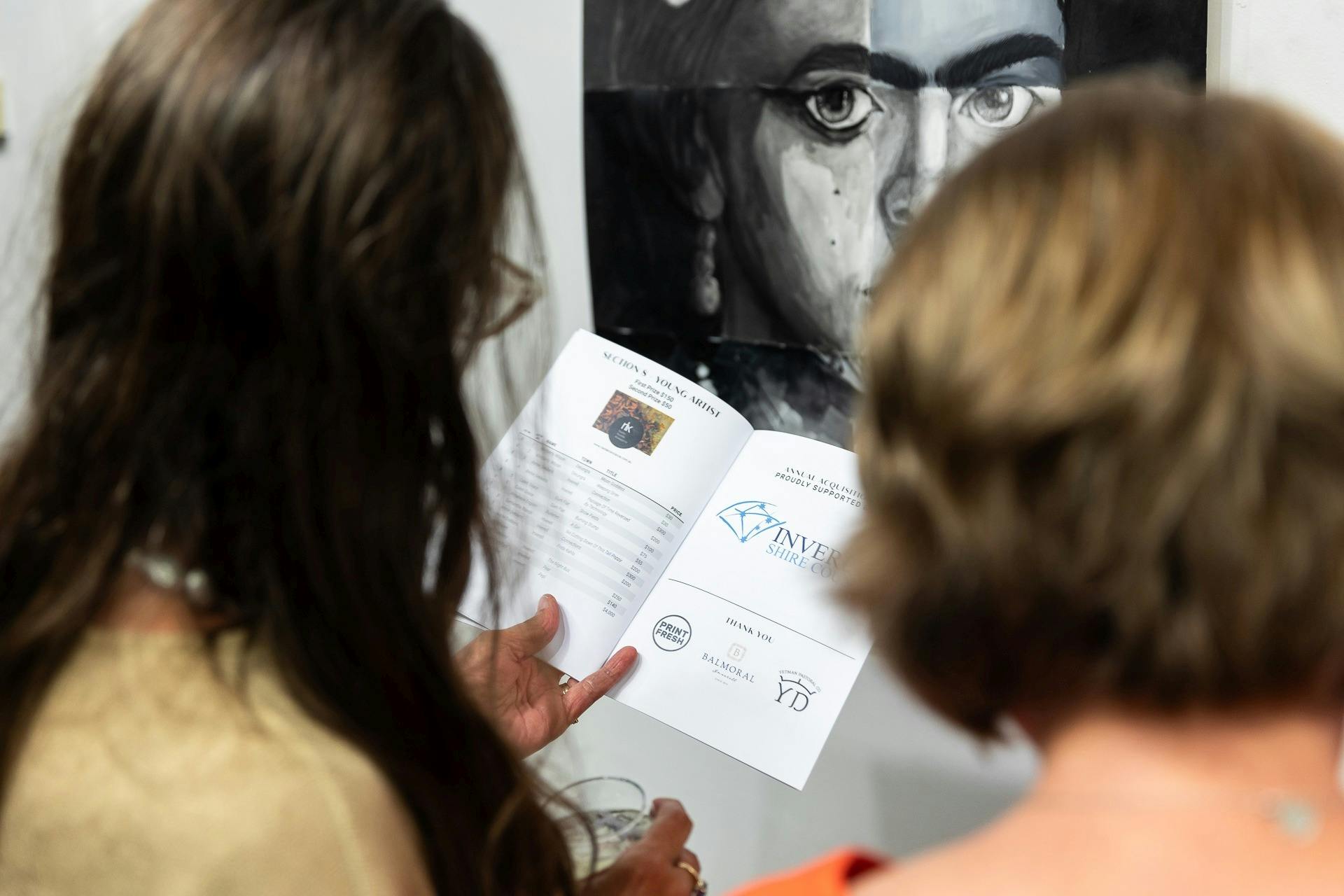 Back of 2 woman looking at a catalogue of exhibitions while standing in front of a portrait