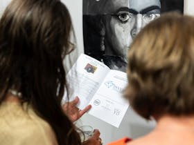 Back of 2 woman looking at a catalogue of exhibitions while standing in front of a portrait