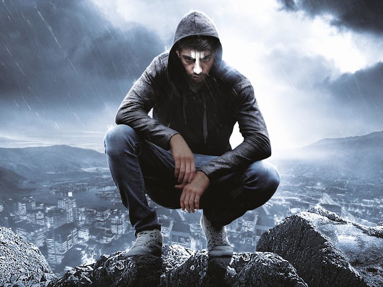 Person crouched on top of rocks wearing a hoodie and face paint with dark clouds and a city behind