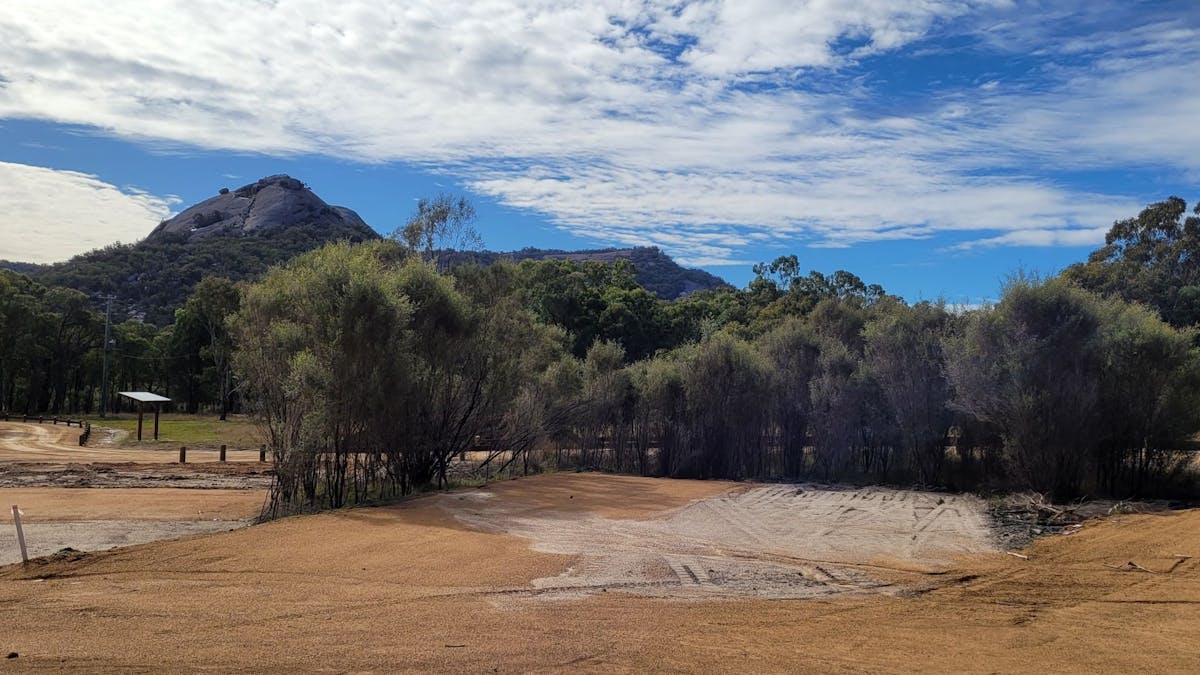 A large dirt camp site surround by native trees. A granite rock formation is in the background.
