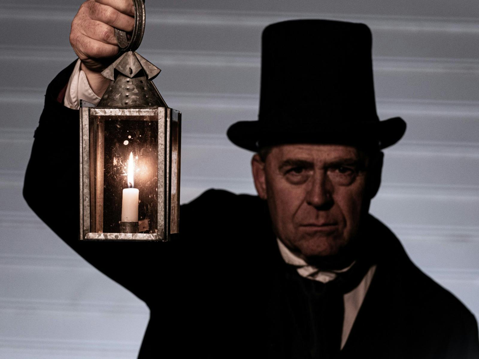 Man dressed in black jacket, white shirt and top hat holding up a lantern