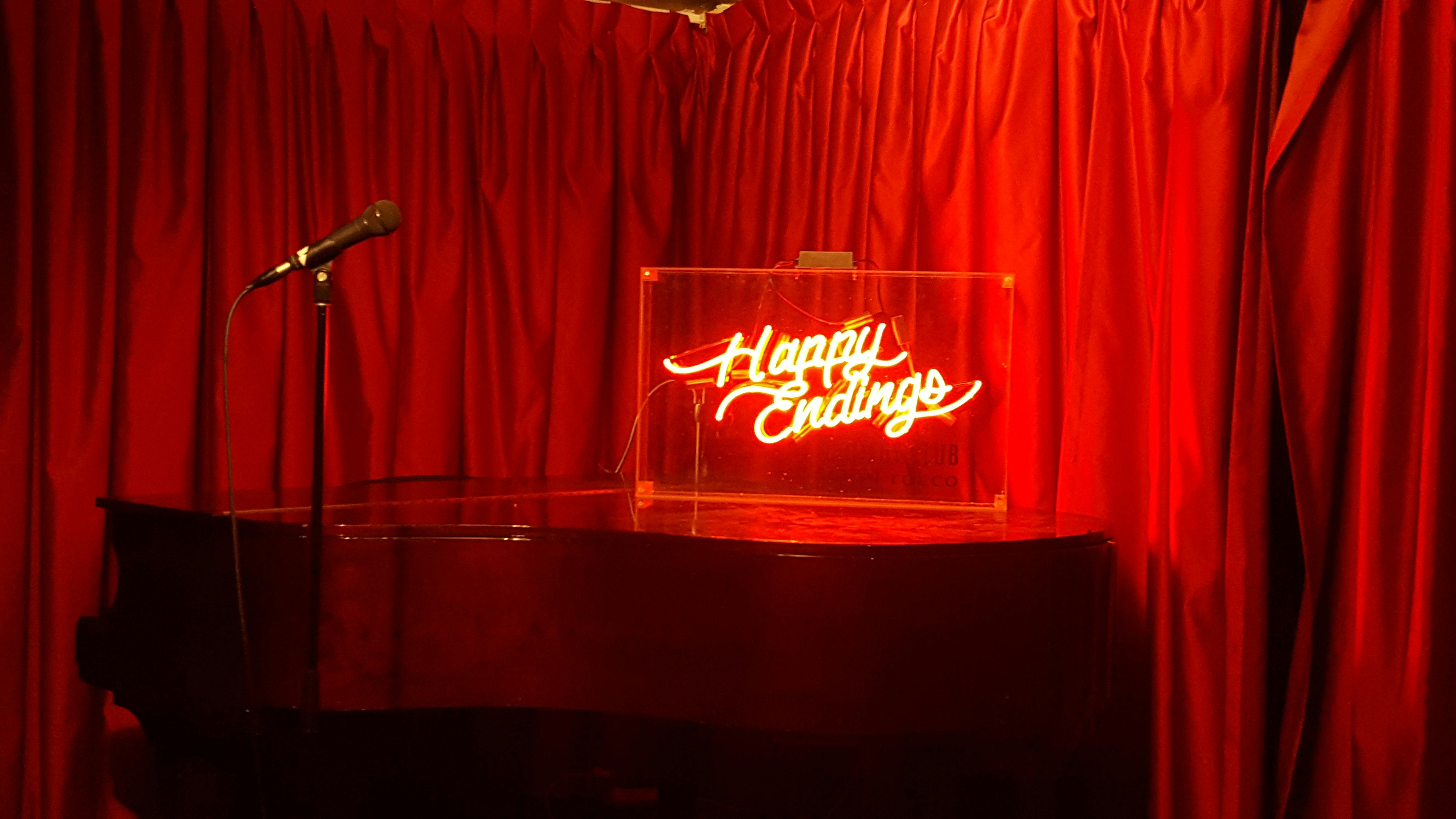 Microphone with neon-lit sign reading 'Happy Endings', with a red velvet curtain back drop.