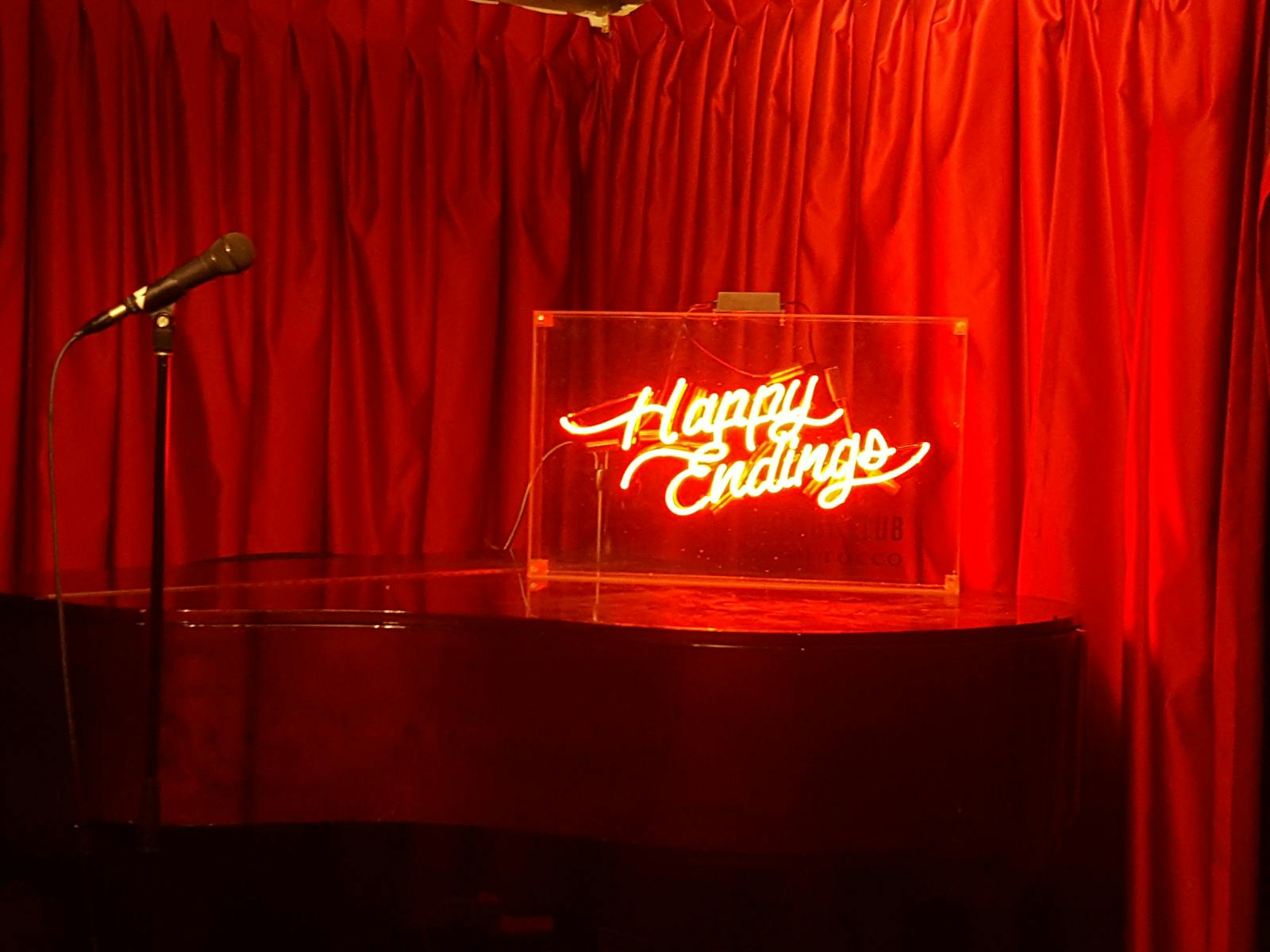 Microphone with neon-lit sign reading 'Happy Endings', with a red velvet curtain back drop.