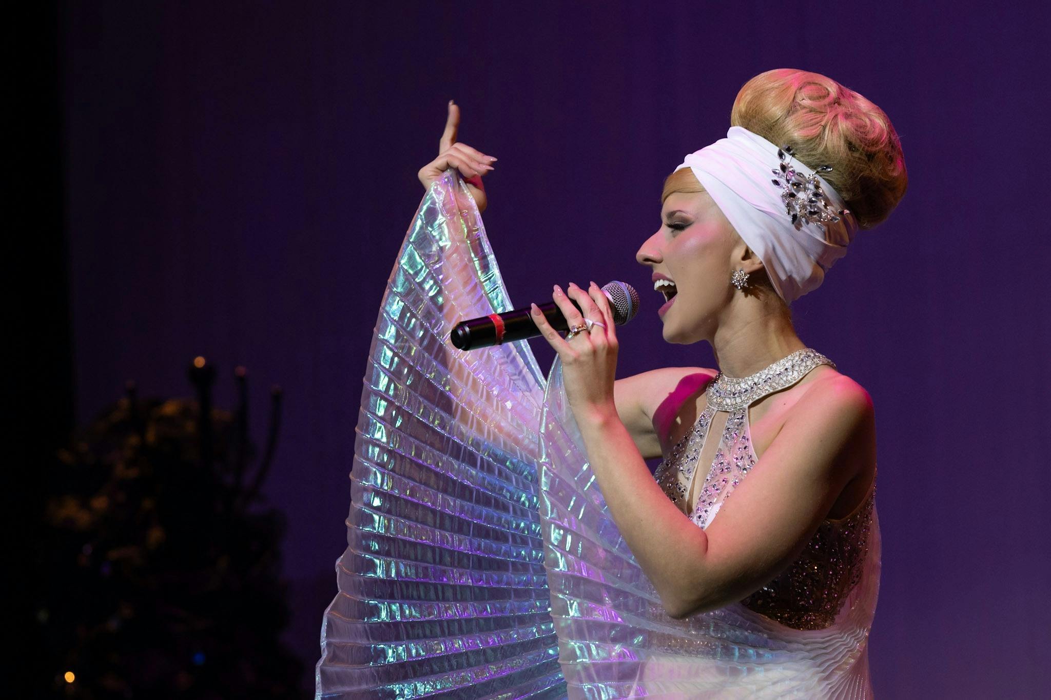 Rachael Grace, impersonating Barbra Streisand, in glamorous 60s clothes, sings on stage.