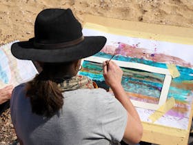 A woman in a wide brim hat is sitting and painting in watercolour while sitting on sand.