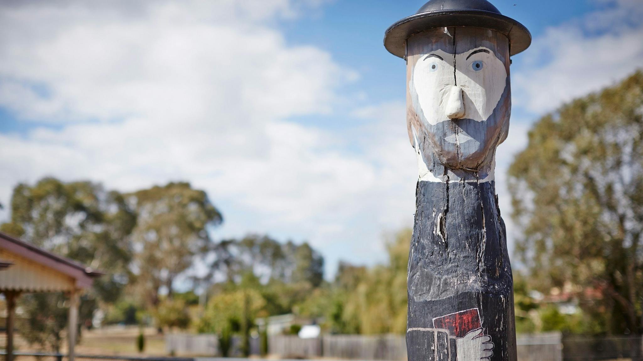 Bollard that looks like a bearded man, wearing a hat, and holding a book, trees, clouds, sunny day.