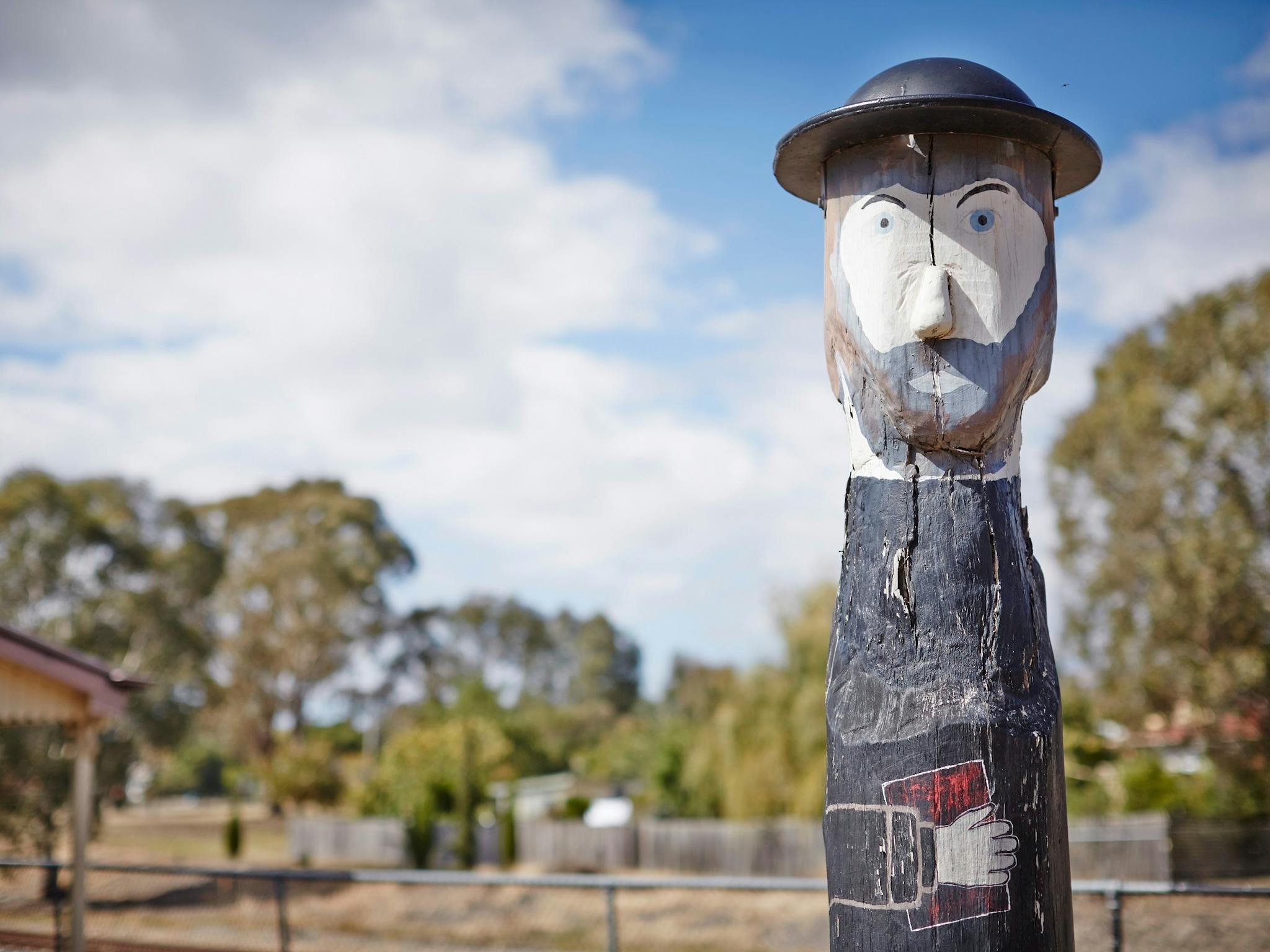 Bollard that looks like a bearded man, wearing a hat, and holding a book, trees, clouds, sunny day.