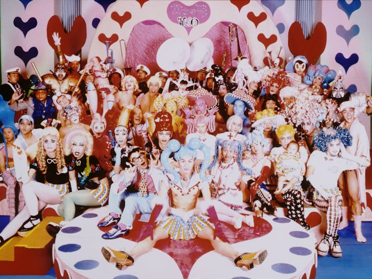 A large group of people in bright costumes on a colourful stage decorated with love hearts.