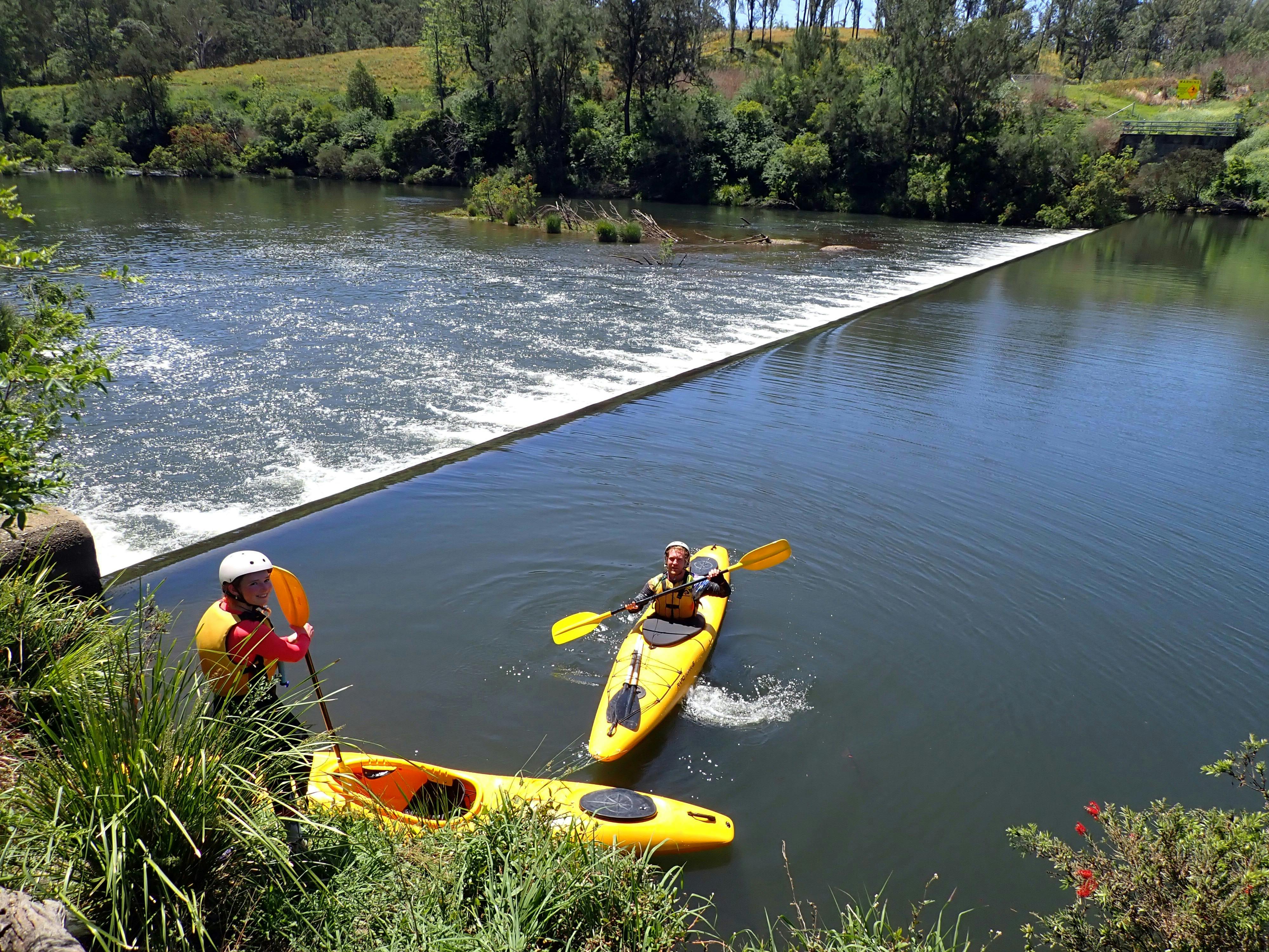 A person is sitting in a kayak with water flowing over the  weir  behind  the kayaks