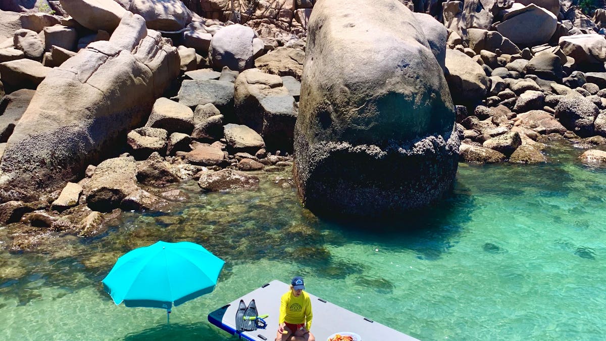 lady sitting on a raft in water eating morning tea near the beach with a child swimming next to her