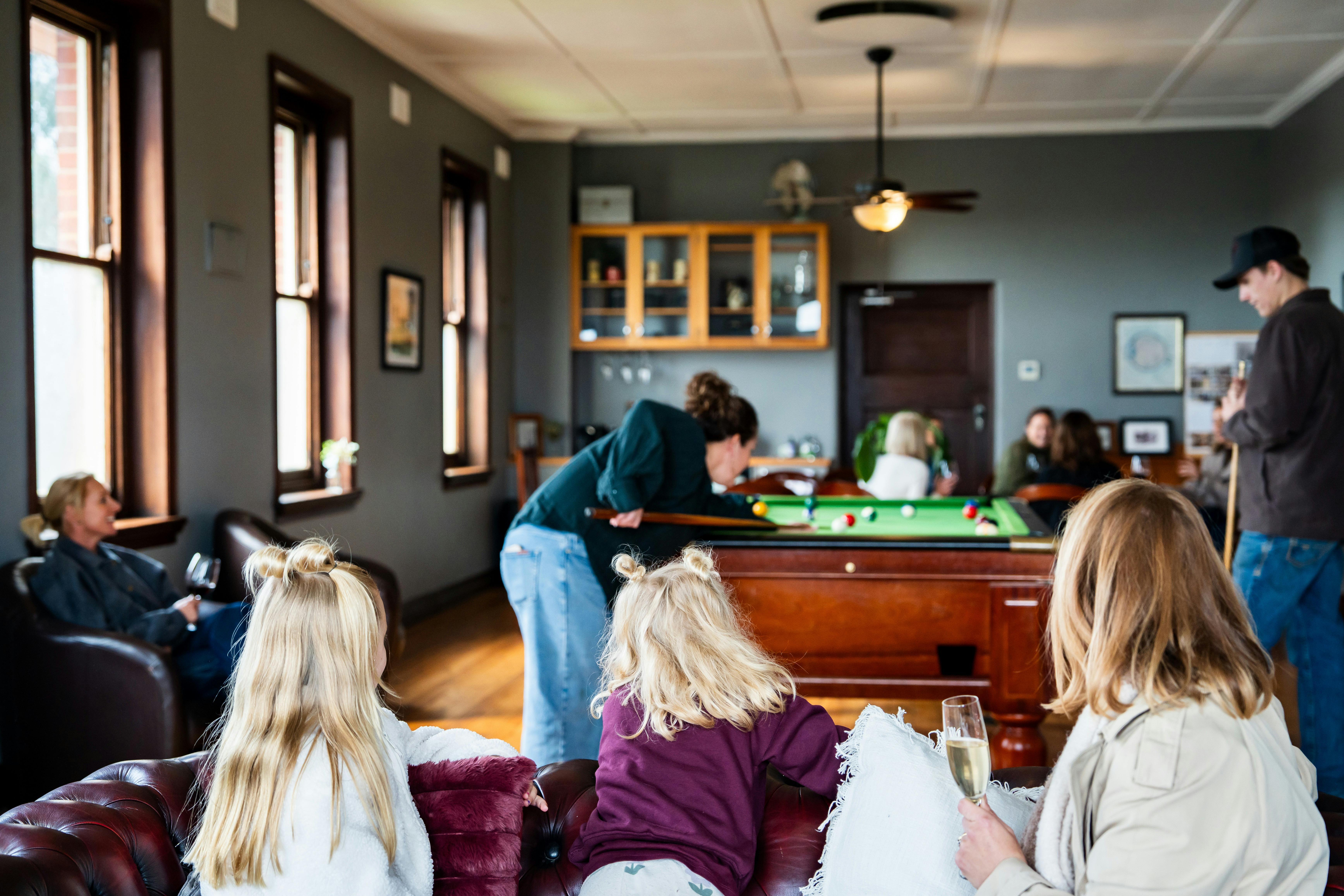 Kids sitting on couch watching some adults playing pool