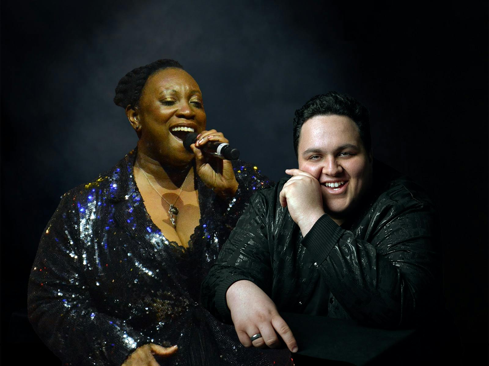 A woman in a shiny dress and a man in dark clothing smile at the camera, the woman holding a mic