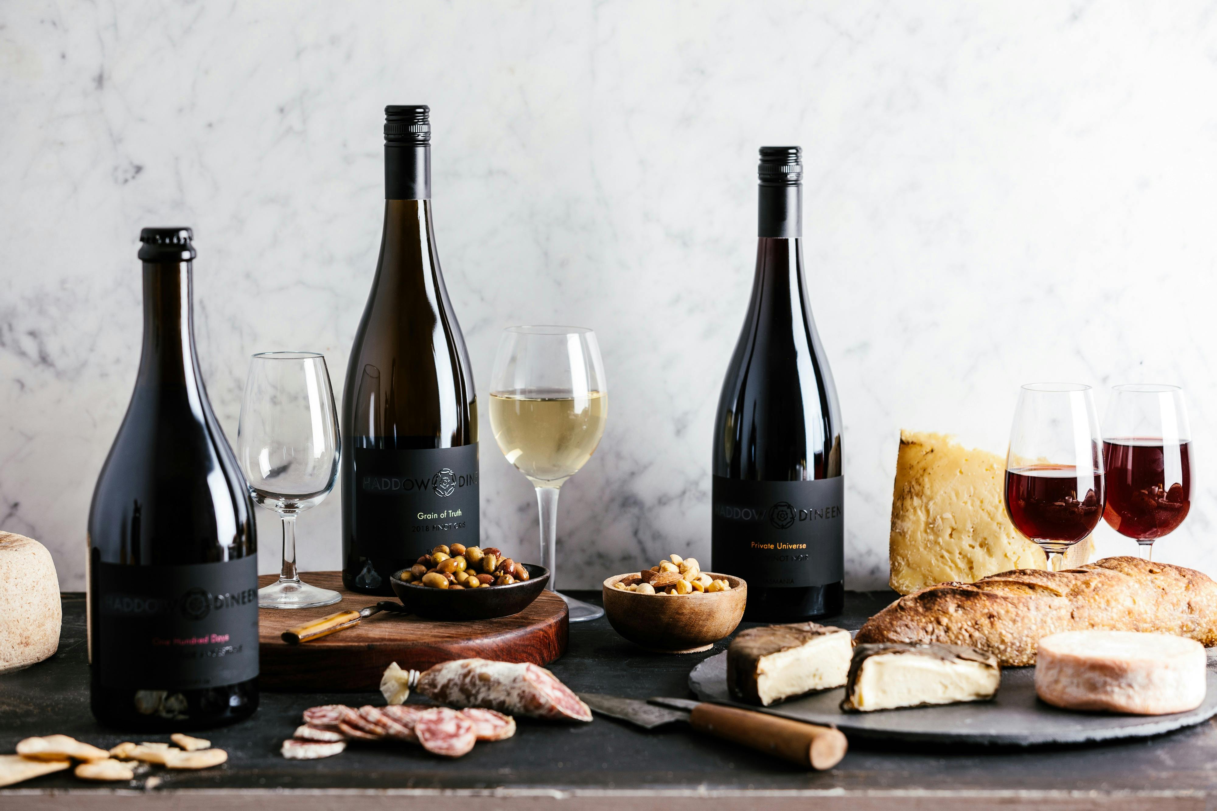 three bottles of wine placed alongside glassware, cheese, and meat items