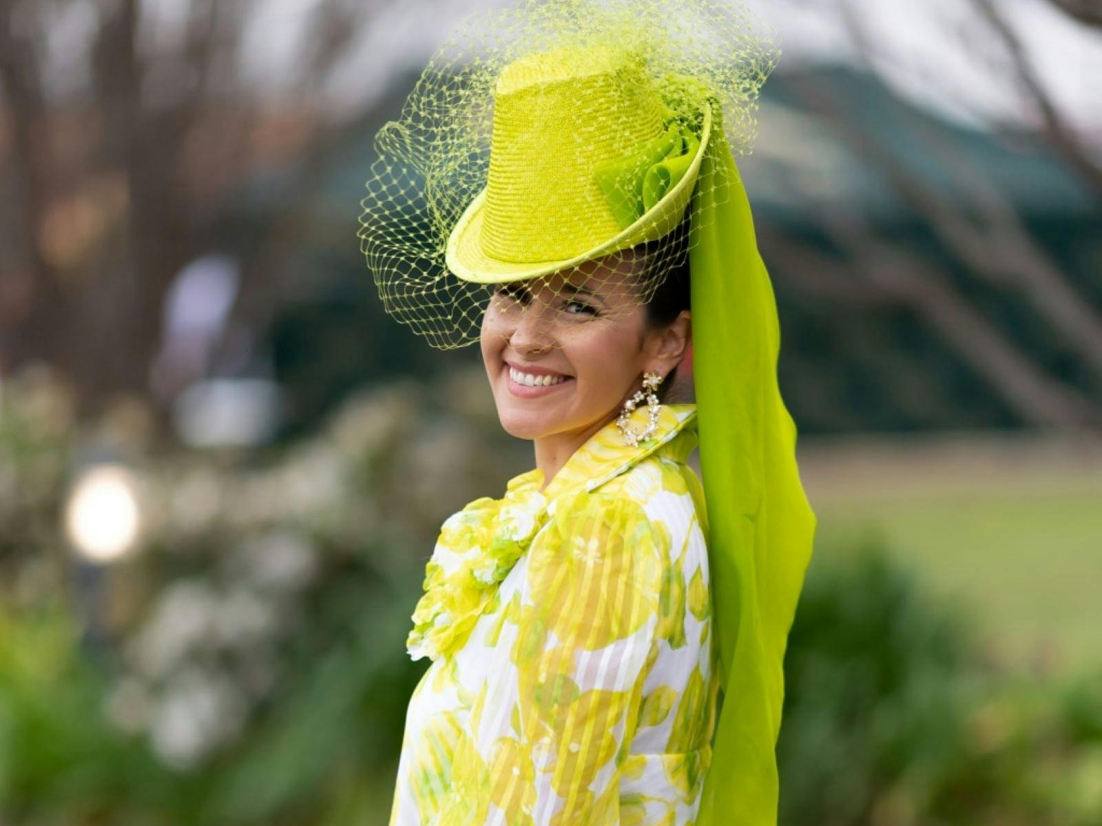 A woman with a bright green top hap fascinator and veil wearing a fashionable white and yellow top