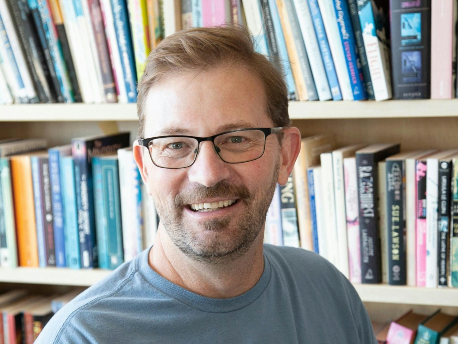 Man with glasses smiles in front of bookshelf