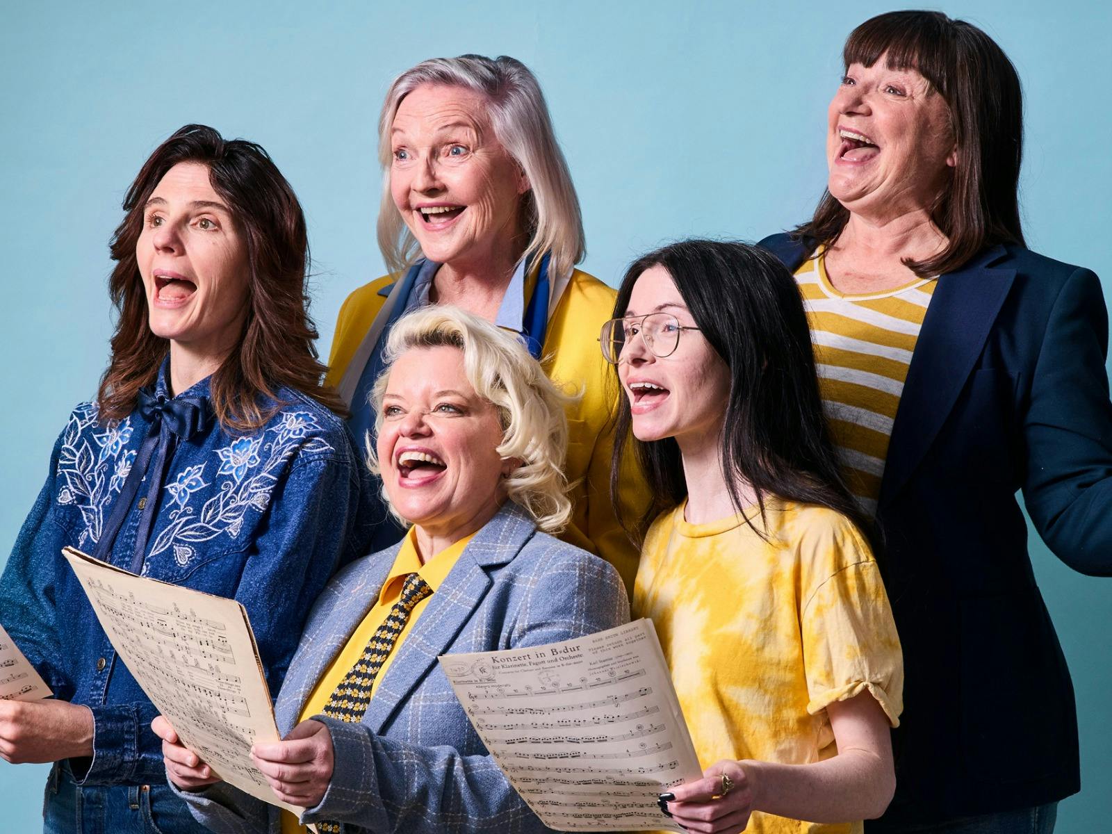Five women in colorful casual clothes stand together, laughing and singing from sheet music against