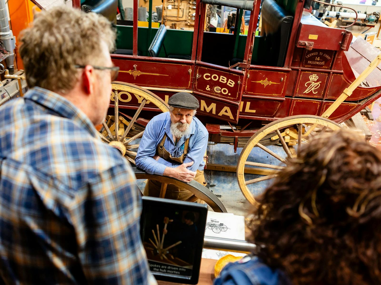 2 museum visitors discuss wheelwrighting with craftsman. 2 museum visitors discuss wheelwrighting with craftsman.