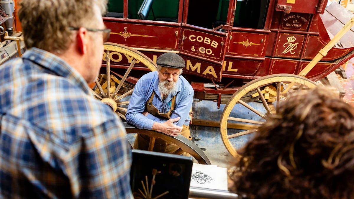 2 museum visitors discuss wheelwrighting with craftsman.