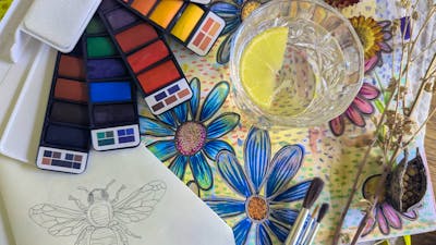 Watercolour palletes with painted flowers and a sketched bee next to a gin and tonic.