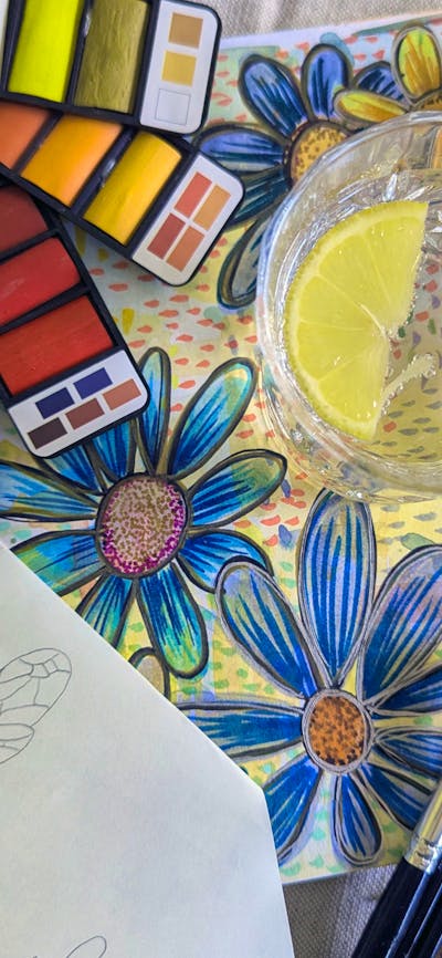Watercolour palletes with painted flowers and a sketched bee next to a gin and tonic.
